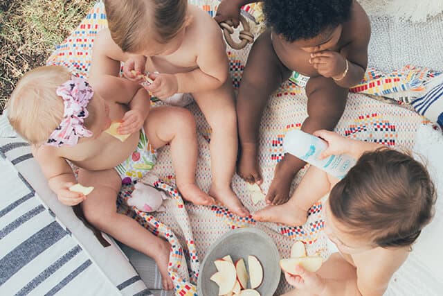 Four babies sitting on a blanket eating snacks